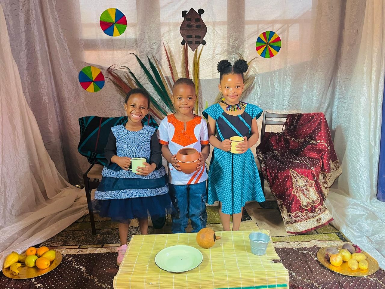 Three children in traditional attire holding a clay pot and enamel mugs on the Heritage Day set